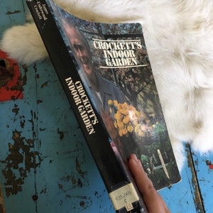 Puede incluir: Un libro en blanco y negro titulado "Crockett's Indoor Garden" de James Underwood Crockett. La portada del libro presenta una foto de un hombre con una planta en el fondo.