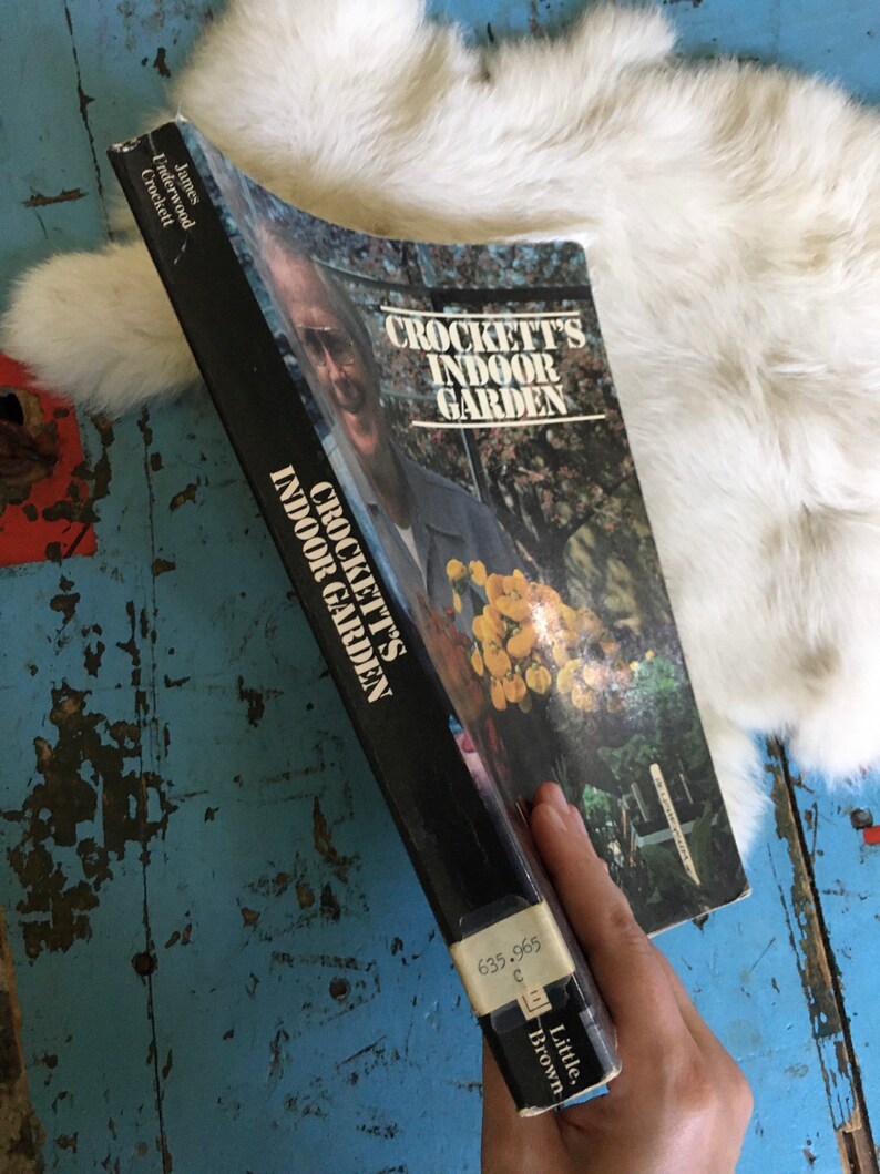 Puede incluir: Un libro en blanco y negro titulado "Crockett's Indoor Garden" de James Underwood Crockett. La portada del libro presenta una foto de un hombre con una planta en el fondo.