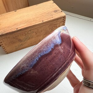 May include: A handmade ceramic bowl with a unique shape and a glossy finish. The bowl features a deep burgundy interior with a blue and white rim. The exterior is a light beige color. A wooden box is in the background.