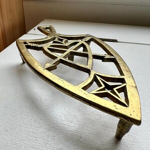 Pode incluir: Um descanso de panela de metal dourado com um design de estrela e recortes intrincados. O descanso de panela tem tr&ecirc;s pernas e tem a forma de um escudo.