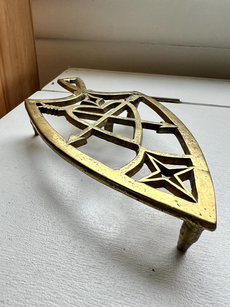 Pode incluir: Um descanso de panela de metal dourado com um design de estrela e recortes intrincados. O descanso de panela tem tr&ecirc;s pernas e tem a forma de um escudo.