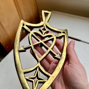 Pode incluir: Um descanso de panela de metal dourado em forma de escudo com um design de cora&ccedil;&atilde;o e flechas. O descanso de panela tem um recorte em forma de estrela na parte inferior.