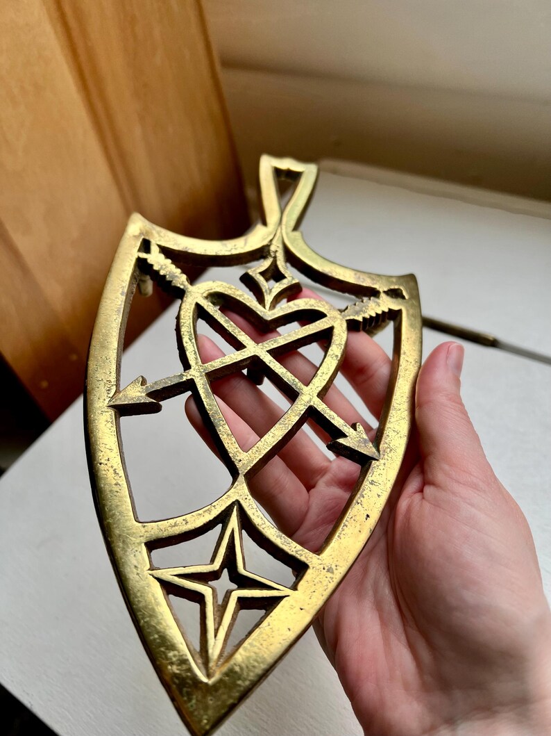 Pode incluir: Um descanso de panela de metal dourado em forma de escudo com um design de cora&ccedil;&atilde;o e flechas. O descanso de panela tem um recorte em forma de estrela na parte inferior.