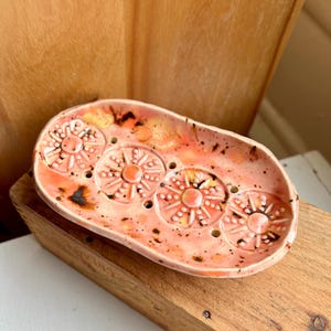 May include: An oval ceramic soap dish with a glossy peach and orange glaze featuring four embossed sunburst patterns. The dish has a raised rim and small drainage holes positioned between the circular sun motifs, with dark brown speckles.