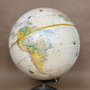 Może przedstawiać: Antyczny globus z białym tłem i żółtymi, zielonymi i niebieskimi kontynentami. Globus stoi na metalowej podstawie z ozdobną podstawą.