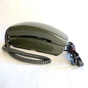 May include: A dark green rotary phone with a coiled cord. The phone has a simple design with a black receiver and a black dial.