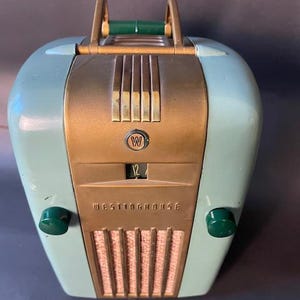 May include: Vintage Westinghouse radio in a rounded, two-tone design. The face is a bronze color, with the word "Westinghouse" and a dial. The sides are a light blue-green, with green knobs. A handle arches over the top.