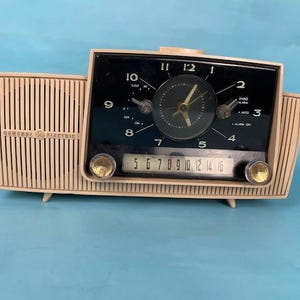 May include: A vintage General Electric clock radio with a cream-colored plastic case and a black face. The clock face has a black background with white numbers and hands. The radio dial has a black background with white numbers. The radio has a sleep timer, alarm, and auto functions.
