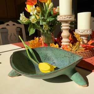 May include: A green ceramic bowl with a unique shape and a yellow tulip inside. The bowl has a speckled glaze and two legs on each side. The bowl is sitting on a white table with a red tablecloth in the background.