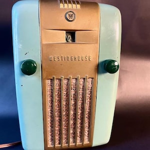 May include: Vintage Westinghouse radio in a teal and gold color scheme. The radio has a rectangular shape with rounded edges and two green knobs. The front features the Westinghouse logo and the brand name.