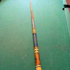 May include: A wooden pool cue featuring a light brown tapered shaft, a textured silver-colored wrap section, and a dark blue leather-like grip. The butt end displays alternating light and dark wood segments with black rings and a cream-colored base.
