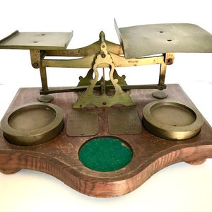 May include: Antique brass postal scale with two weighing platforms and a wooden base. The scale has a green felt pad in the center of the base.