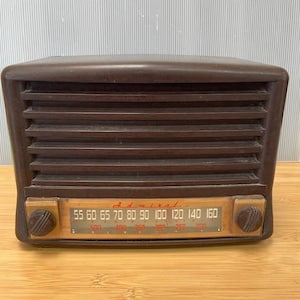 May include: A vintage brown Admiral radio with a horizontal speaker grill and a wooden faceplate. The faceplate displays radio frequency numbers and two knobs. The radio sits on a wooden surface.