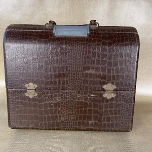 May include: A vintage brown faux-crocodile skin suitcase with a handle and metal clasp. The suitcase has a rectangular shape with rounded edges and decorative metal hardware. The textured surface gives it a classic, retro aesthetic.