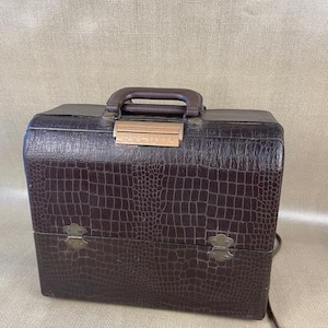 May include: A vintage brown faux-crocodile skin makeup case with a handle and metal clasps. The case has a rectangular shape and a hinged lid. The case has a brown handle and a light brown clasp. A brown electrical cord is attached to the side.