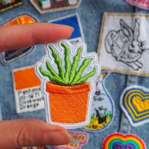 May include: A green succulent plant in a small orange pot, embroidered on a white fabric patch. The patch is held by a finger against a denim background.