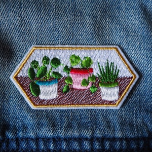 May include: A white embroidered patch with a hexagonal shape features three potted plants. The plants are green and have white pots with brown details. The patch has a gold outline.
