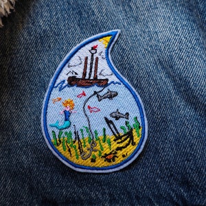 May include: A blue and yellow embroidered patch depicting a mermaid swimming in the ocean with a ship above. The patch is shaped like a teardrop and has a white outline.
