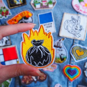 Pode incluir: Um saco de lixo preto com chamas bordadas em um fundo amarelo. O remendo é segurado por uma mão contra um fundo de jeans com outros remendos coloridos.