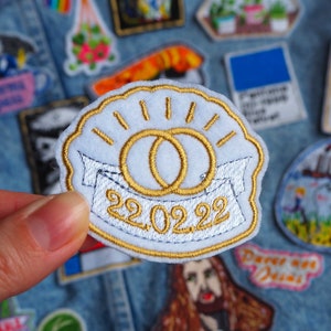 Puede incluir: Un parche de fieltro blanco con un diseño bordado en oro de dos anillos entrelazados y la fecha 22.02.22. El parche está rodeado de otros parches sobre un fondo de mezclilla.