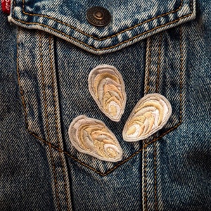 Op de afbeelding: Close-up van een spijkerjasje met drie geborduurde oesterpatches in crème en bruin. De jas heeft een zak met een metalen knoop en zichtbare stiksels.