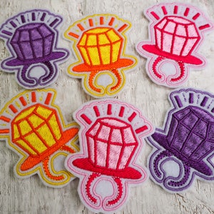 May include: Six embroidered iron-on patches featuring a ring with a diamond in the centre. The rings are in different colours: purple, yellow, pink, and red. The diamonds are outlined in white.