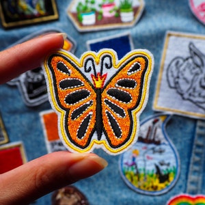 May include: Orange and black embroidered butterfly patch with a crown on its head. The patch is on a blue denim background.