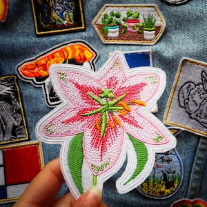 Pode incluir: Um remendo branco bordado de um lírio rosa com folhas verdes. O remendo está sobre um fundo de ganga com outros remendos.