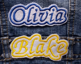 May include: Two embroidered patches on a blue denim jacket. The top patch is white with blue stitching and says "Olivia". The bottom patch is white with yellow stitching and says "Blake".