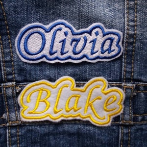 May include: Two embroidered patches on a blue denim jacket. The top patch is white with blue stitching and says "Olivia". The bottom patch is white with yellow stitching and says "Blake".