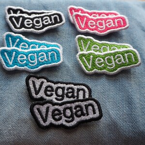 May include: A set of nine iron-on patches with the word "Vegan" in different colours and fonts. The patches are on a blue denim background.