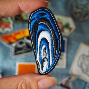 以下が含まれることがあります： アゲートの形をした青い、白と黒の刺繍パッチ。パッチは、層状の同心円状のデザインになっています。