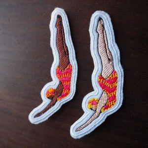 May include: Two embroidered iron-on patches of divers in red, yellow, and brown swimsuits. The patches are on a white background.