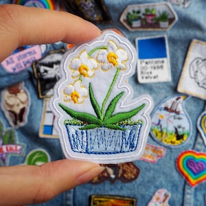 May include: White embroidered flower patch with yellow centres in a blue and white pot. The patch is on a denim background.