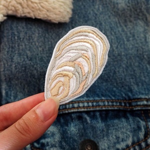 Peut inclure: Un patch brodé blanc en forme de coquille d'huître avec des détails beige et brun clair.