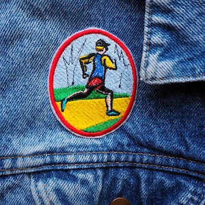 May include: A blue denim jacket with a round embroidered patch featuring a person running. The patch has a red border, a yellow background, and green grass.