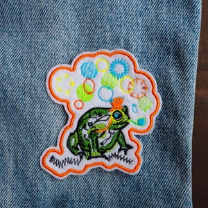 May include: A green frog with a blue eye and orange accents is blowing bubbles. The bubbles are white with colourful outlines. The frog is embroidered on a white patch with an orange border. The patch is sewn onto a blue denim fabric.
