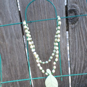 May include: A green and white beaded necklace with a green leaf pendant hanging from a green metal fence.