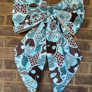 May include: A large fabric bow featuring a quilted texture and a repeating pattern of dark brown, light blue, and white swirling shapes and geometric floral motifs. The bow has two long, pointed tails hanging downward and a central knot.