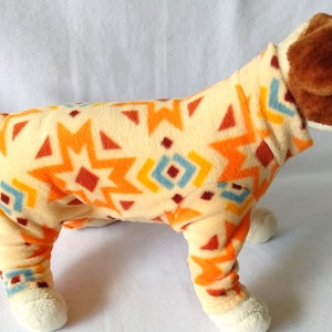 May include: A beige dog pajamas with a geometric pattern in orange, red, blue, and yellow. The pajamas have a turtleneck and long legs.