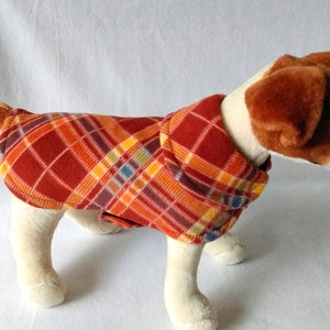 May include: A red, orange, and yellow plaid fleece dog sweater. The sweater has a fold-over collar and is worn by a small, white and brown dog.