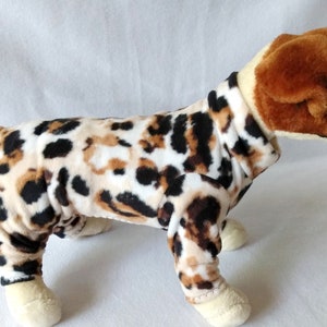 May include: A plush dog toy wearing a leopard print outfit. The dog has a brown and white head and tail, with cream-colored paws. The outfit covers the dog's body in a pattern of black and brown spots on a cream background.