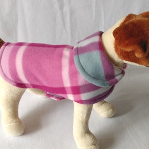 May include: A pink and blue plaid fleece dog coat with a white trim. The coat is designed for a small dog.