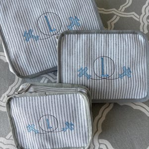 May include: Set of three gray and white striped fabric items. Each item features a blue embroidered letter "L" inside a circle, with a blue airplane design. The edges are trimmed with gray fabric.