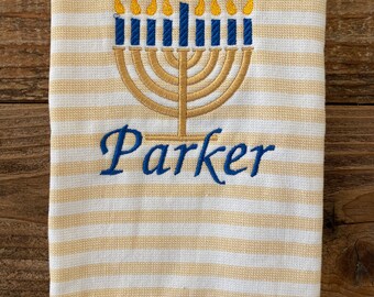 May include: A white and tan striped towel with an embroidered menorah design. The menorah is gold with blue candles and yellow flames. The name "Parker" is embroidered in blue script below the menorah.