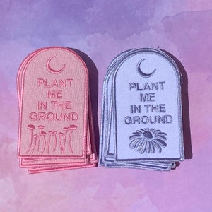 May include: Two stacks of fabric patches with the text "Plant Me In The Ground" and a crescent moon. One stack is pink with mushrooms and the other is blue with a daisy.