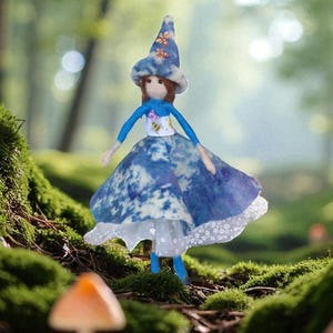 Könnte beinhalten: Handgefertigte Puppe mit blau-weißem Kleid und Hut, verziert mit floralen Akzenten. Die Puppe hat braunes Haar und blaue Gliedmaßen. Die Puppe steht in einer Waldszene mit Moos und Pilzen.