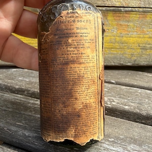 May include: An antique, dark glass bottle with a worn, aged paper label. The label features faded text in German, with the words "Angostura Bittern" visible. The bottle is held by a hand, suggesting its size and age.