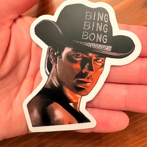 以下が含まれることがあります： 「BING BING BONG」の文字が点線で描かれた黒いカウボーイハットをかぶった人物の肖像画が描かれたステッカー。肖像画は暗い背景にあり、真剣な表情をしています。