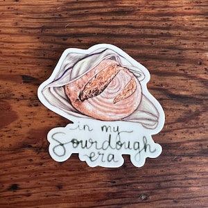 May include: A white cloth-covered sourdough bread loaf with the text "in my sourdough era" written in black cursive.